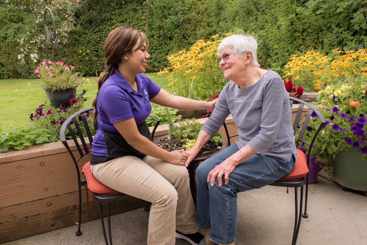 A caregiver interacts with an elderly woman in a garden setting.