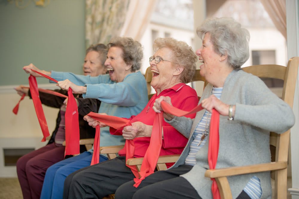A group of elderly women joyfully participating in an exercise activity with red bands while seated on chairs.