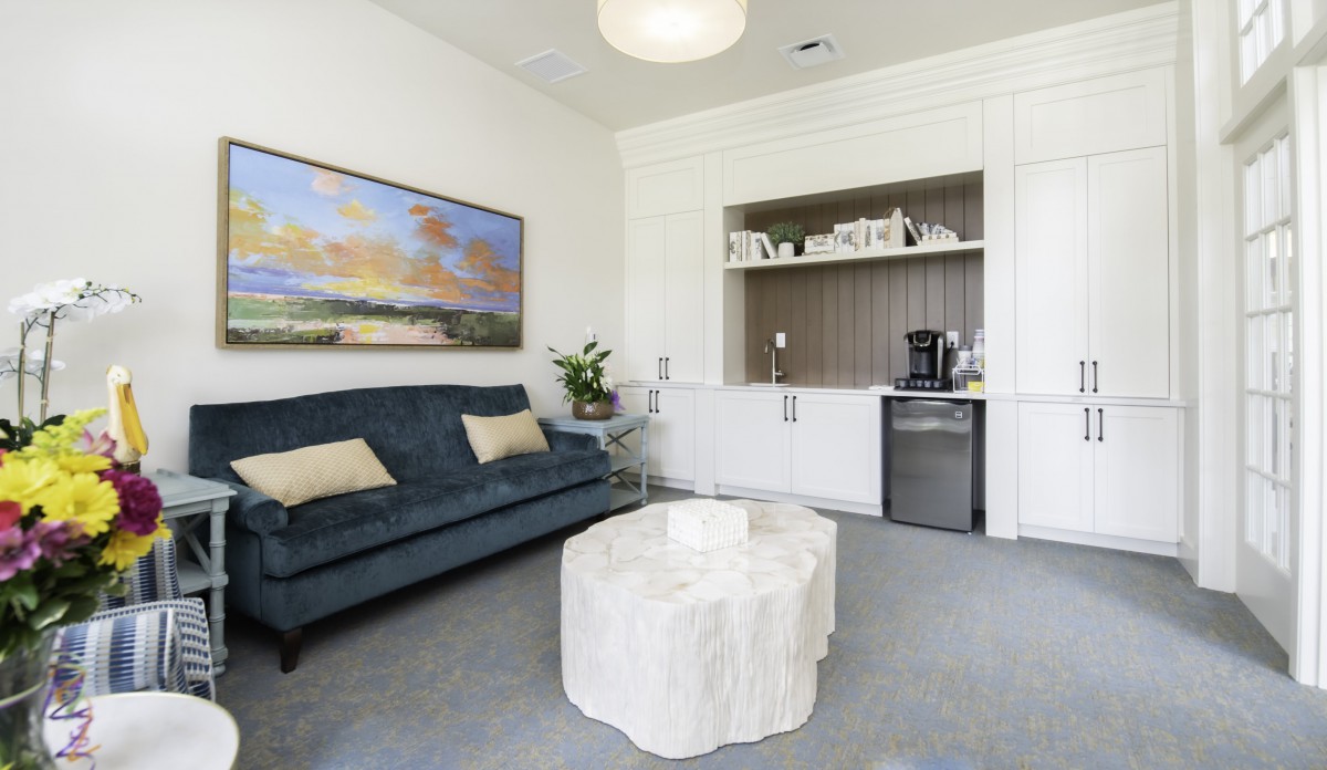 A modern living room with a stylish sofa, unique coffee table, and artistic wall painting. The space includes built-in shelving displaying decorative items, alongside a mini fridge and coffee maker.