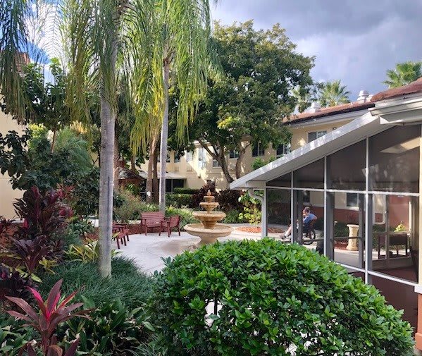 A lush garden patio with a central fountain, surrounded by palm trees and vibrant greenery.