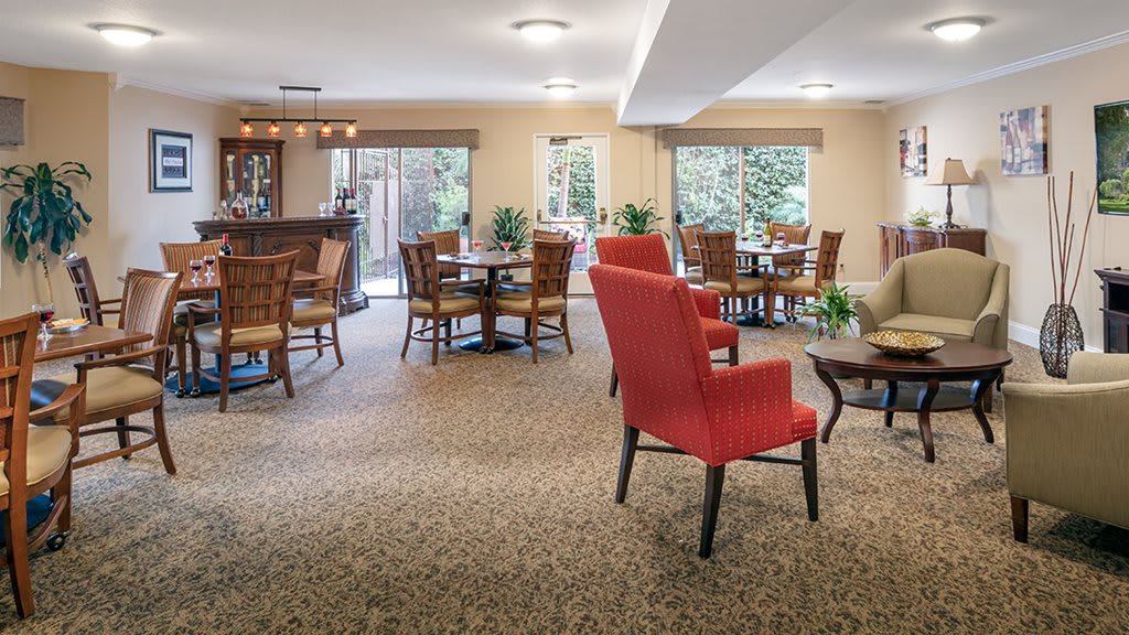 A cozy dining area with tables and red chairs arranged on a carpeted floor, surrounded by large windows and decorative plants.