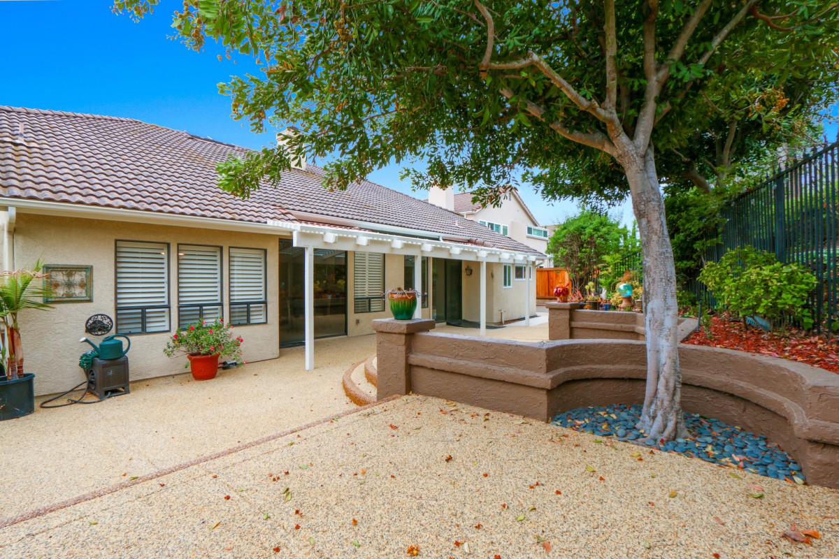 A serene backyard with a tree providing shade over a patio area, featuring a well-maintained garden.