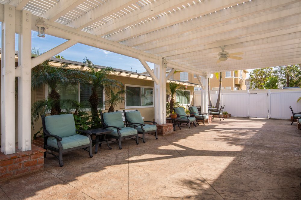 A cozy outdoor patio with cushioned seating under a pergola, surrounded by lush plants on a sunny day.