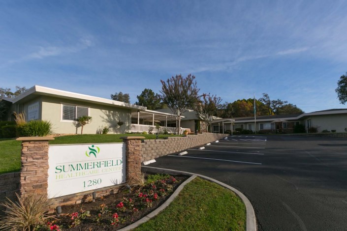 A building with a sign in front, adjacent to a parking lot and surrounded by landscaped greenery.