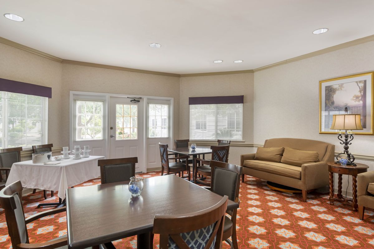 A cozy room with tables, chairs, and a sofa, featuring large windows and a patterned carpet.