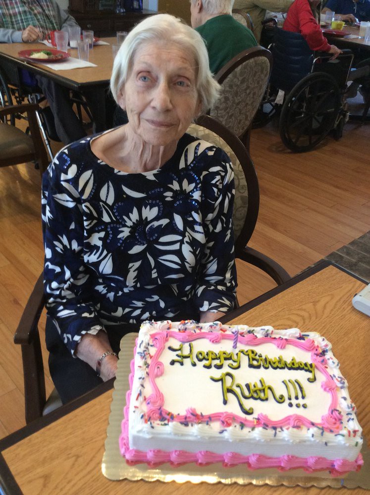 An elderly woman sitting at a table with a birthday cake in front of her.