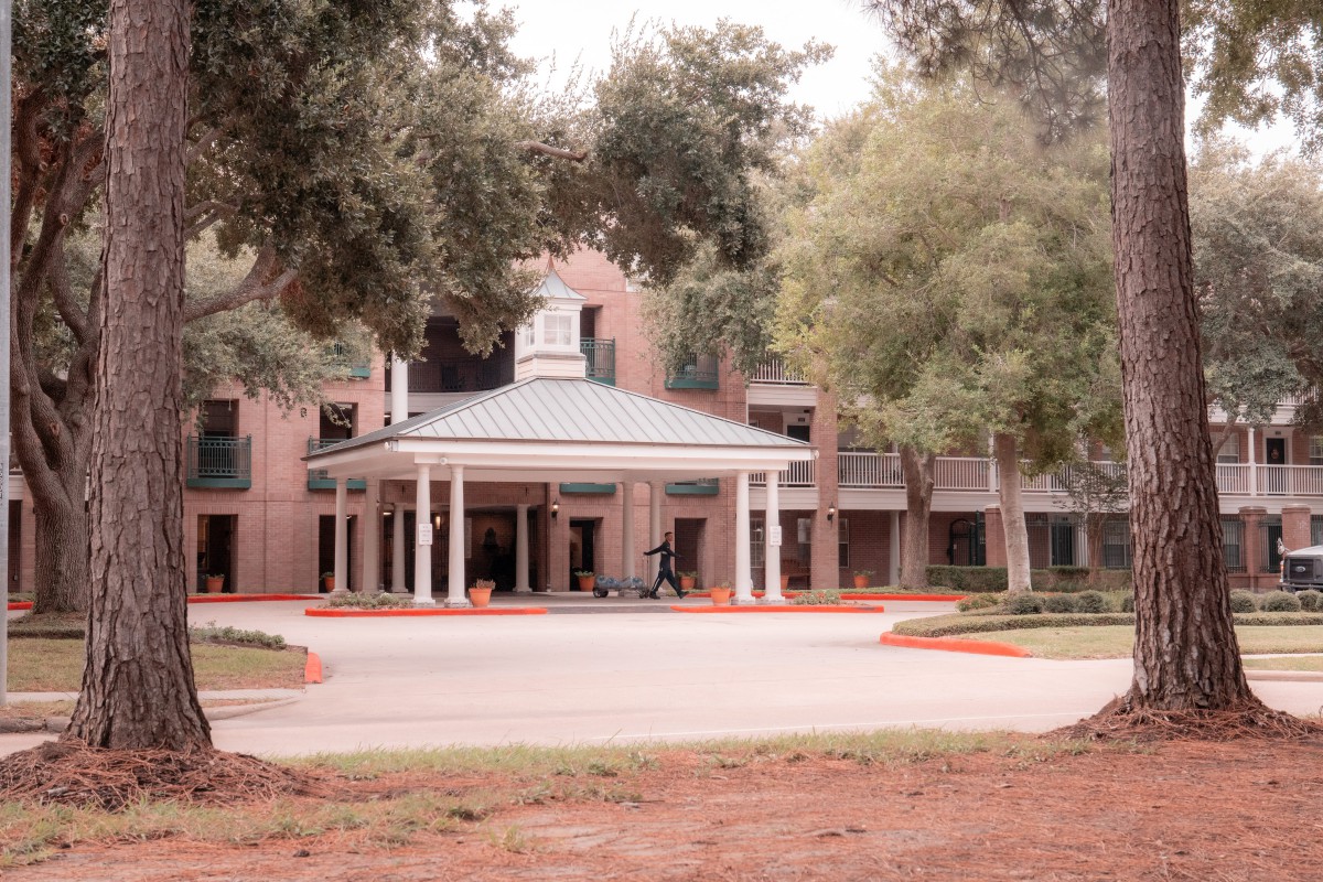 A two-story building with a porch and columns at the entrance, surrounded by trees and a driveway.
