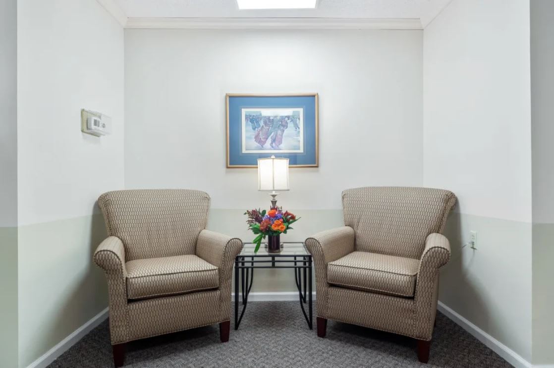 A cozy waiting room with two upholstered chairs, a lamp, and a bouquet of flowers on a table between them.
