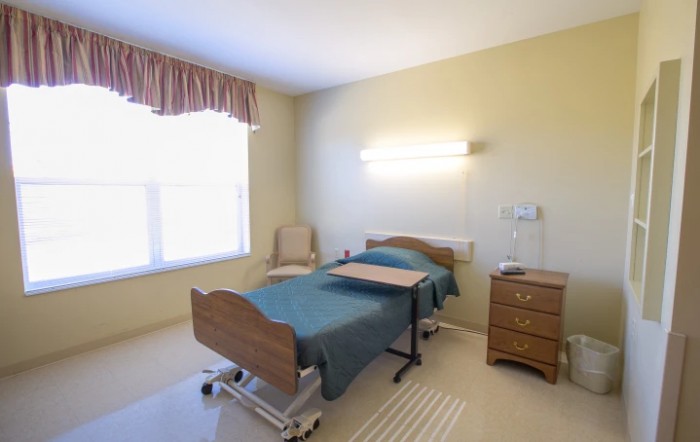 A hospital room with a single bed, bedside table, and a chair next to a large window.