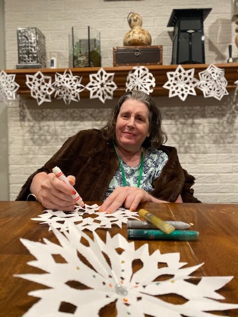A woman is sitting at a table crafting paper snowflakes with a smile on her face.