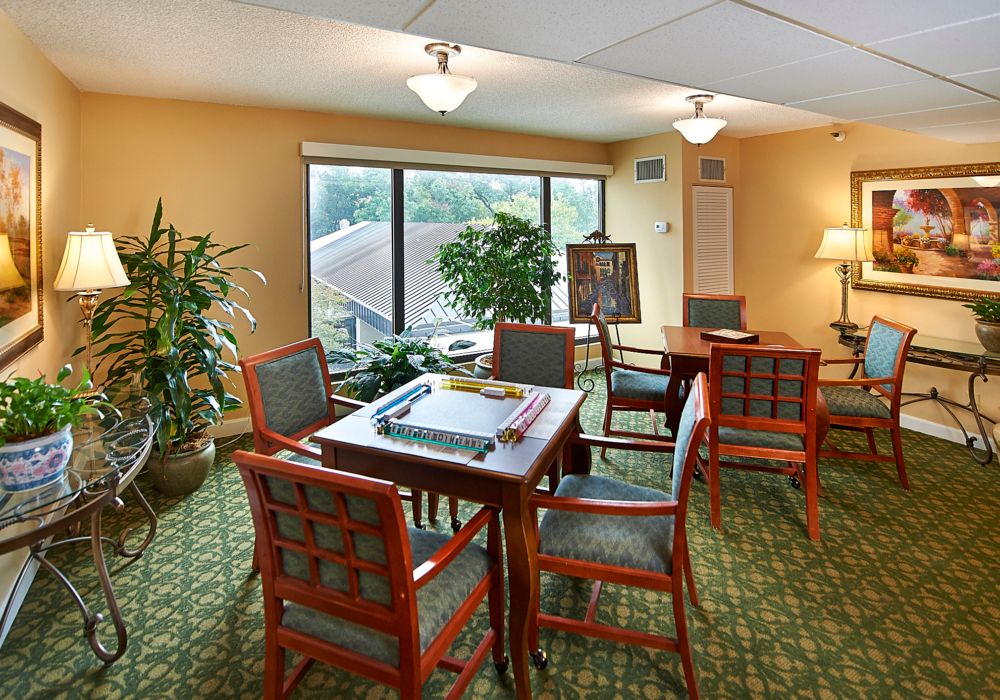 A comfortable, well-lit meeting area with a large window, featuring tables and chairs set for gathering or games, and decorated with plants.