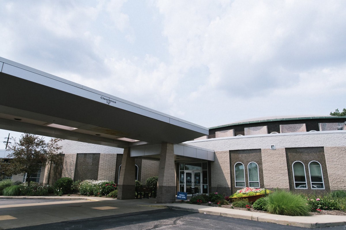 A modern building with a covered entrance and a parking area under a cloudy sky.