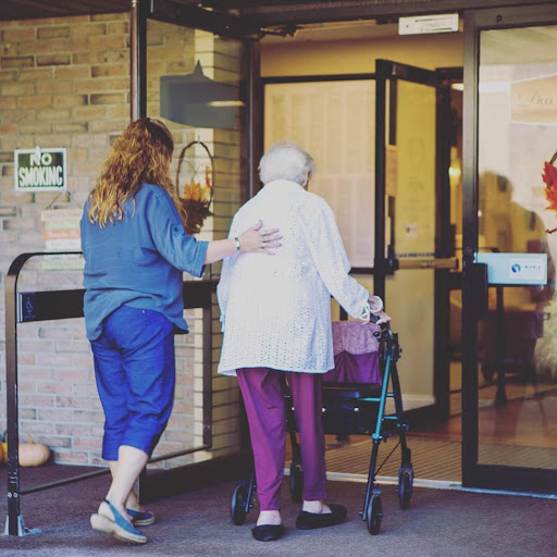 A caregiver assists an elderly woman using a walker as they enter a building.