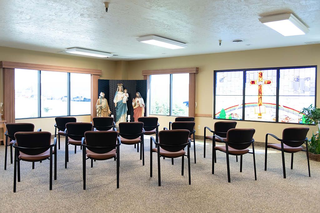 A chapel room with rows of chairs facing religious statues in front of large windows.
