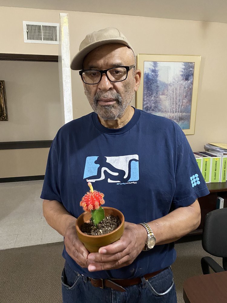 A man wearing a hat and glasses holds a potted plant indoors.