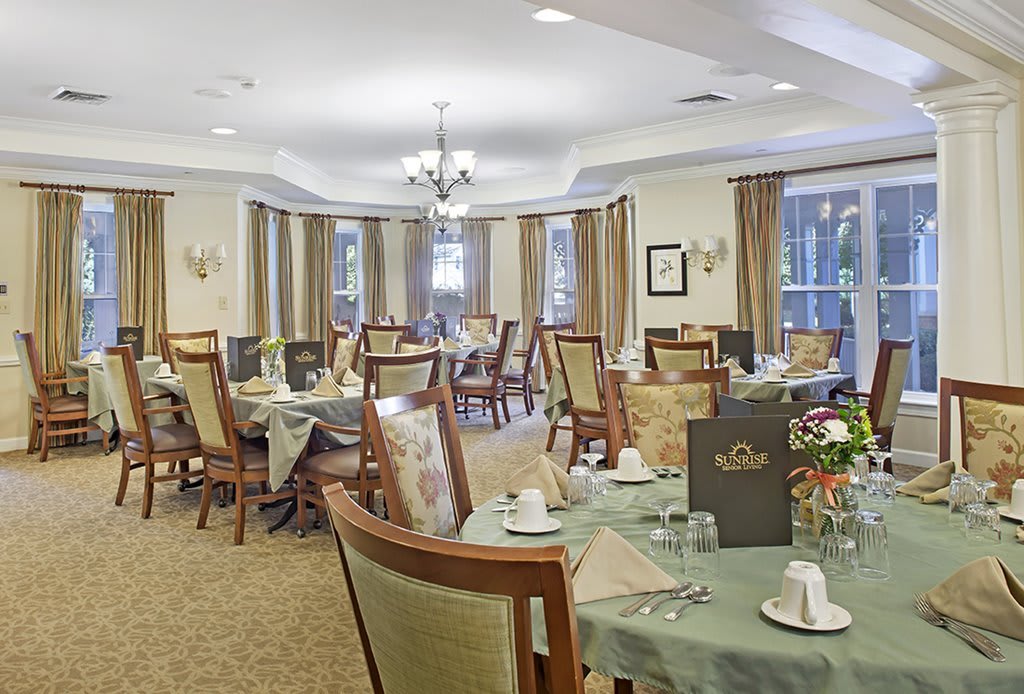 A well-set dining room with neatly arranged tables and chairs, elegant chandeliers, and large windows with curtains.
