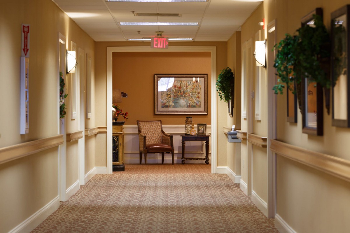 A warmly lit hallway with chairs and a table against a wall decorated with a painting and plants.
