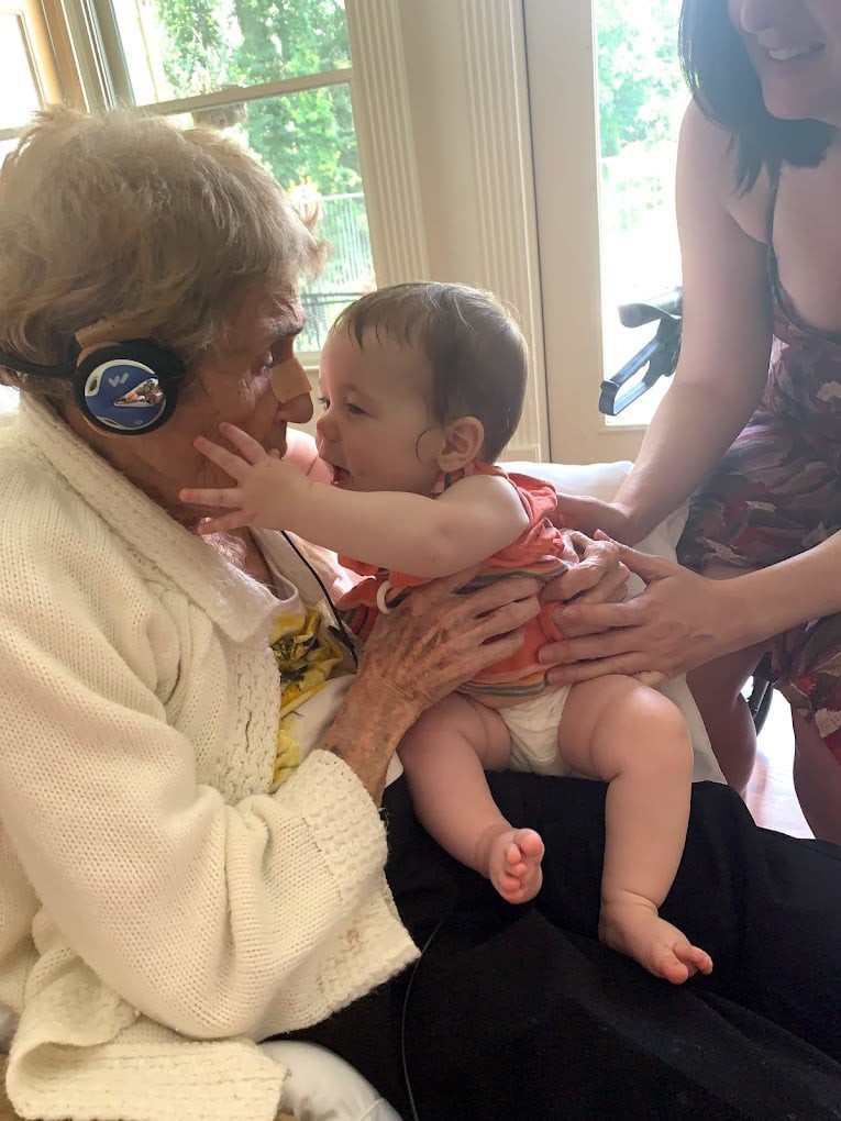 An elderly person wearing headphones shares a tender moment with a baby in their arms, surrounded by family.