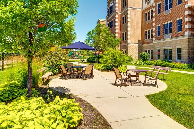 A tranquil outdoor patio area with chairs and a table under an umbrella, surrounded by lush greenery and adjacent to a large building.