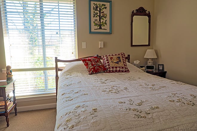 A cozy bedroom featuring a quilt-covered bed with decorative pillows, next to a window with blinds, and furnished with a lamp and mirror.