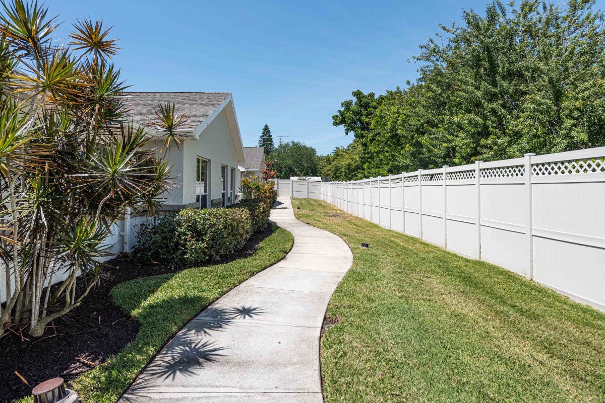 A sunny pathway beside a house with a white fence and green grass.