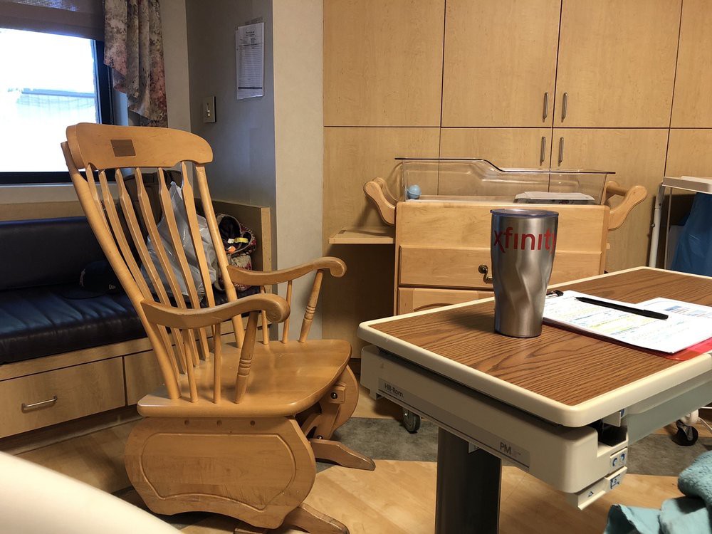 A hospital room featuring a wooden rocking chair, a crib, and a table with a tumbler.