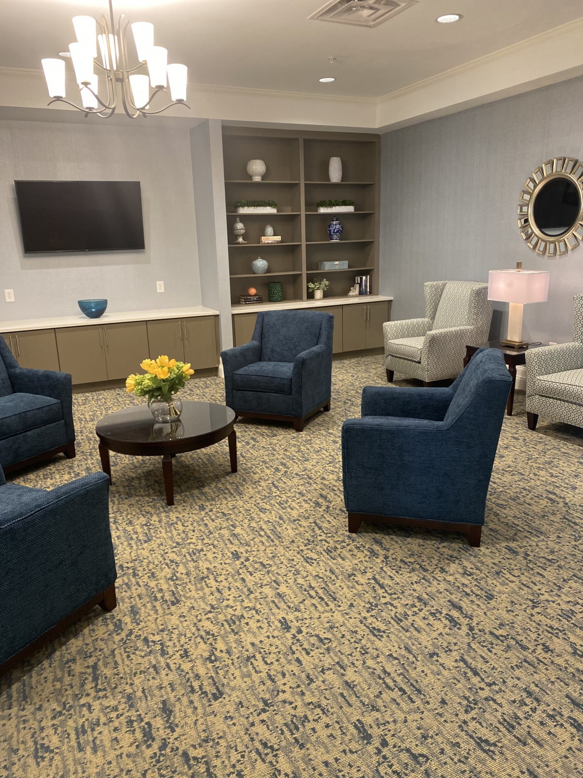 A cozy living room setup with blue and gray chairs, a central table with flowers, a TV on the wall, and decorative shelves.