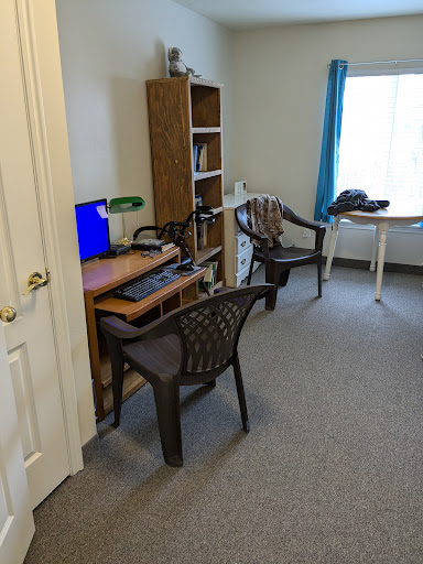 A small office space featuring a desk with a computer, two plastic chairs, and a bookshelf near a window.