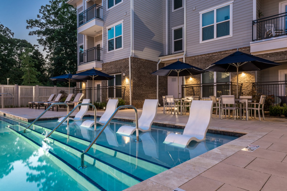 A modern apartment complex features a relaxing swimming pool area with lounge chairs and umbrellas, set against an evening backdrop.
