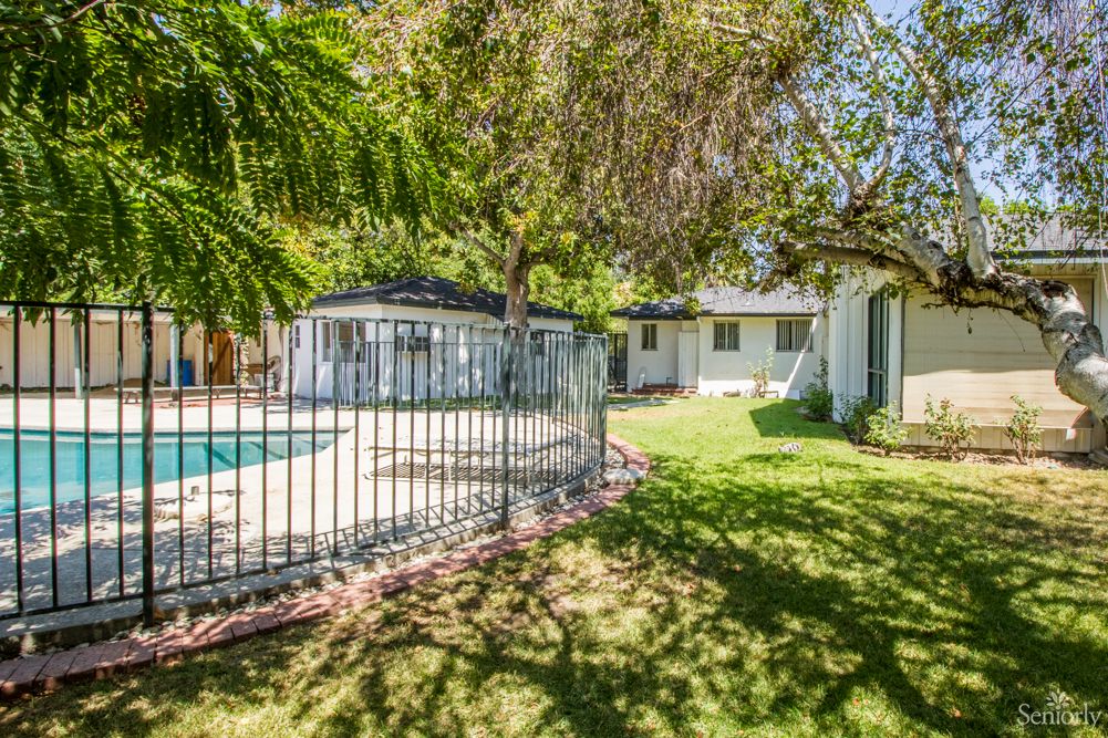 A sunny backyard featuring a pool surrounded by a fence, with lush grass and trees.