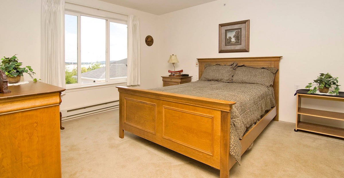 A cozy bedroom with a wooden bed frame, a nightstand with a lamp, and a window allowing natural light.