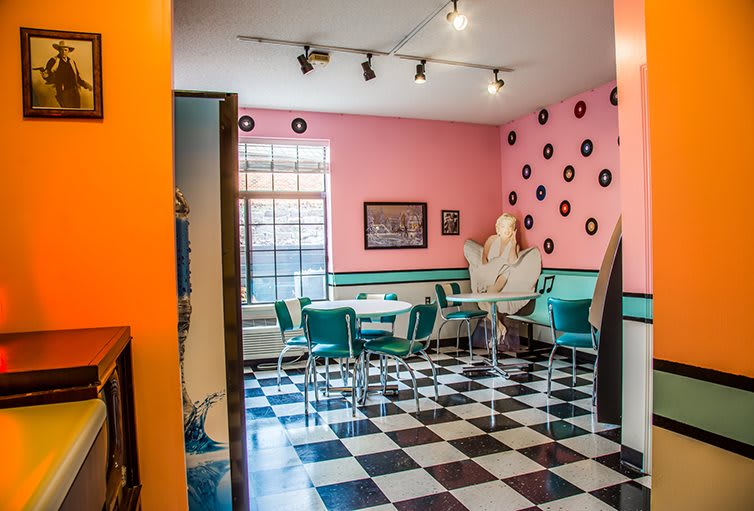 A retro 50s-style diner interior with pink walls, teal chairs, and a checkerboard floor.