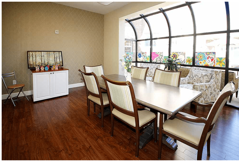 A well-lit dining room with a wooden table and cushioned chairs, featuring a large window and hardwood flooring.