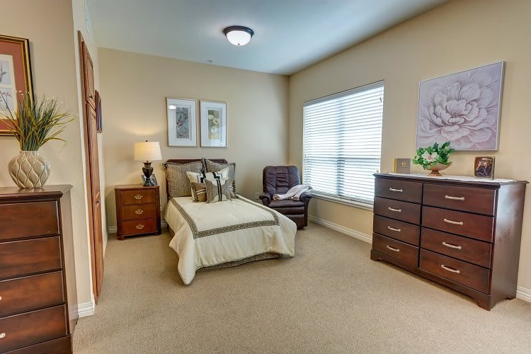 A cozy bedroom with a single bed, an armchair, and a chest of drawers, well-lit by a large window.