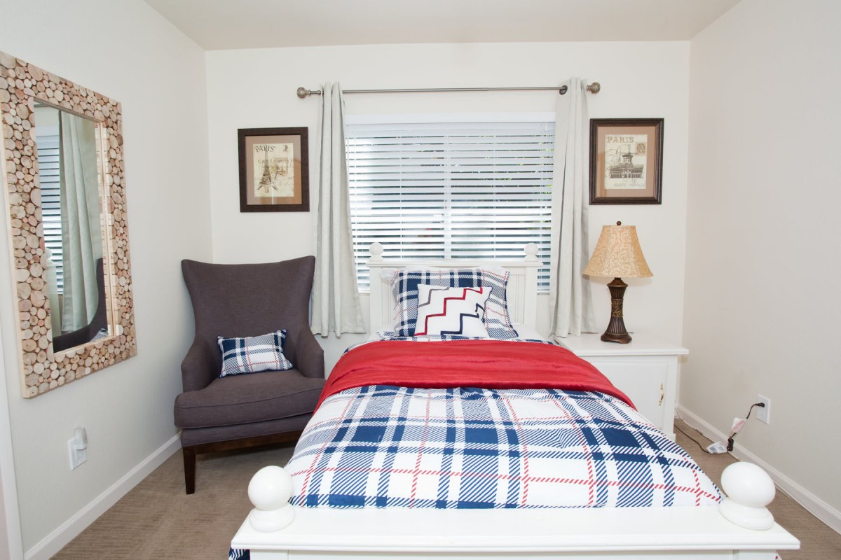A tidy bedroom with a single bed featuring plaid bedding, a chair, and a lamp by the window.