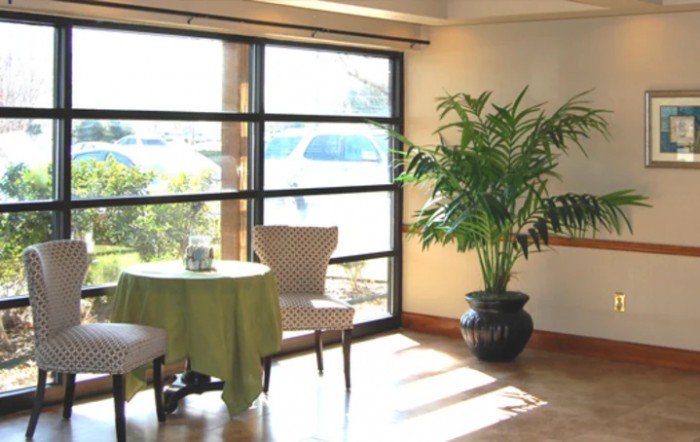 A cozy, sunlit interior with a small table and chairs next to large windows and a potted plant.