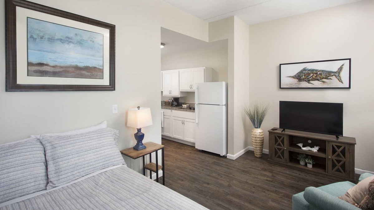 A cozy bedroom with an adjoining kitchenette, featuring a bed, lamp, artwork, and a TV.