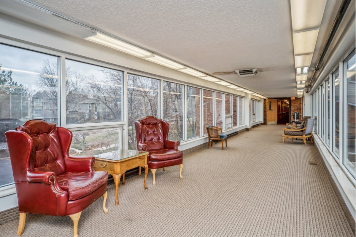 A long hallway with large windows, featuring red leather chairs and a beige carpet.