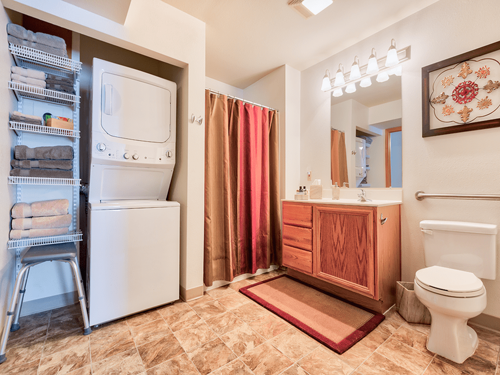 A bathroom with an integrated laundry area, featuring a stacked washer and dryer unit next to a vanity with a large mirror and shower.