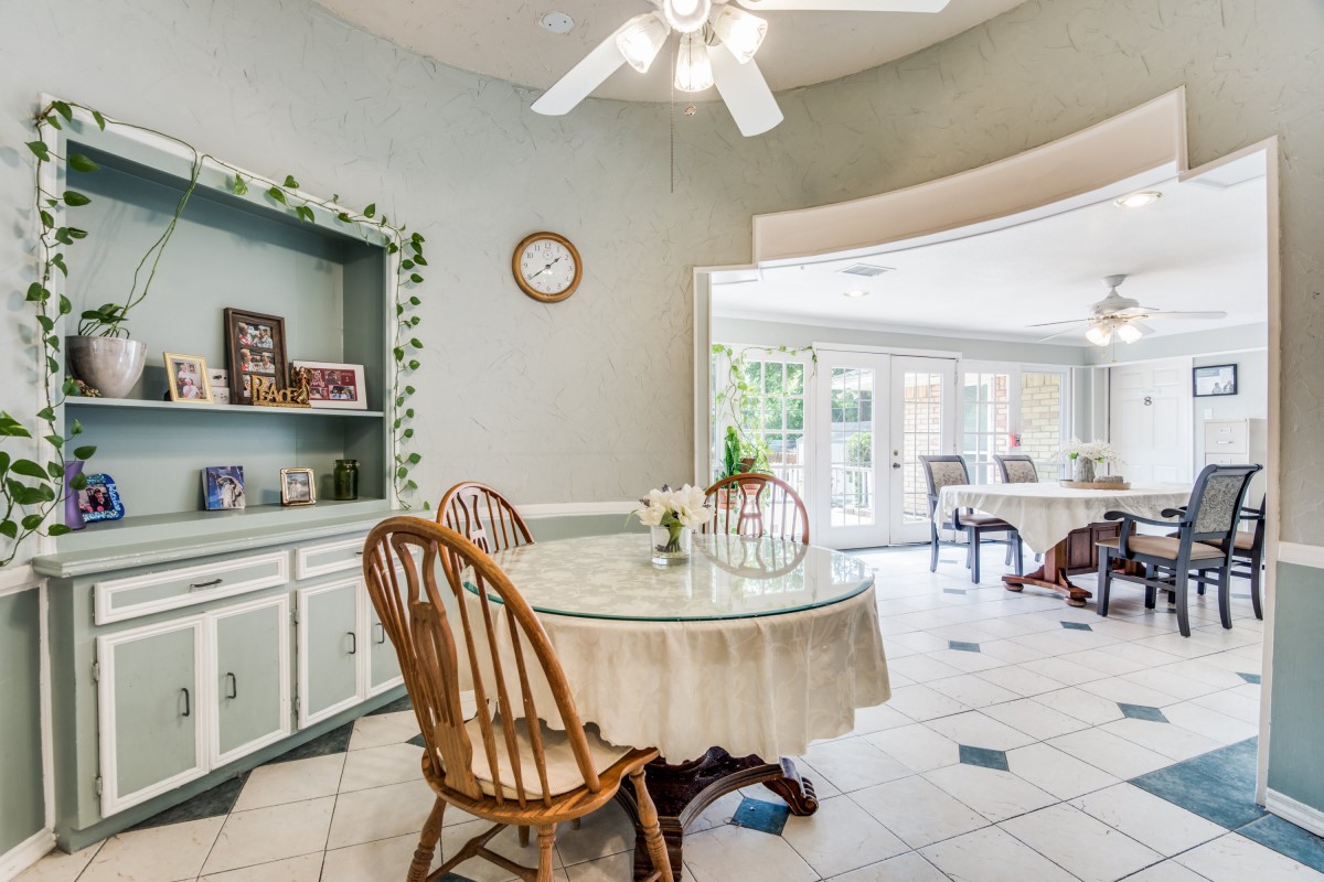 A cozy dining area with a round table and chairs, featuring a ceiling fan and large windows in the background.