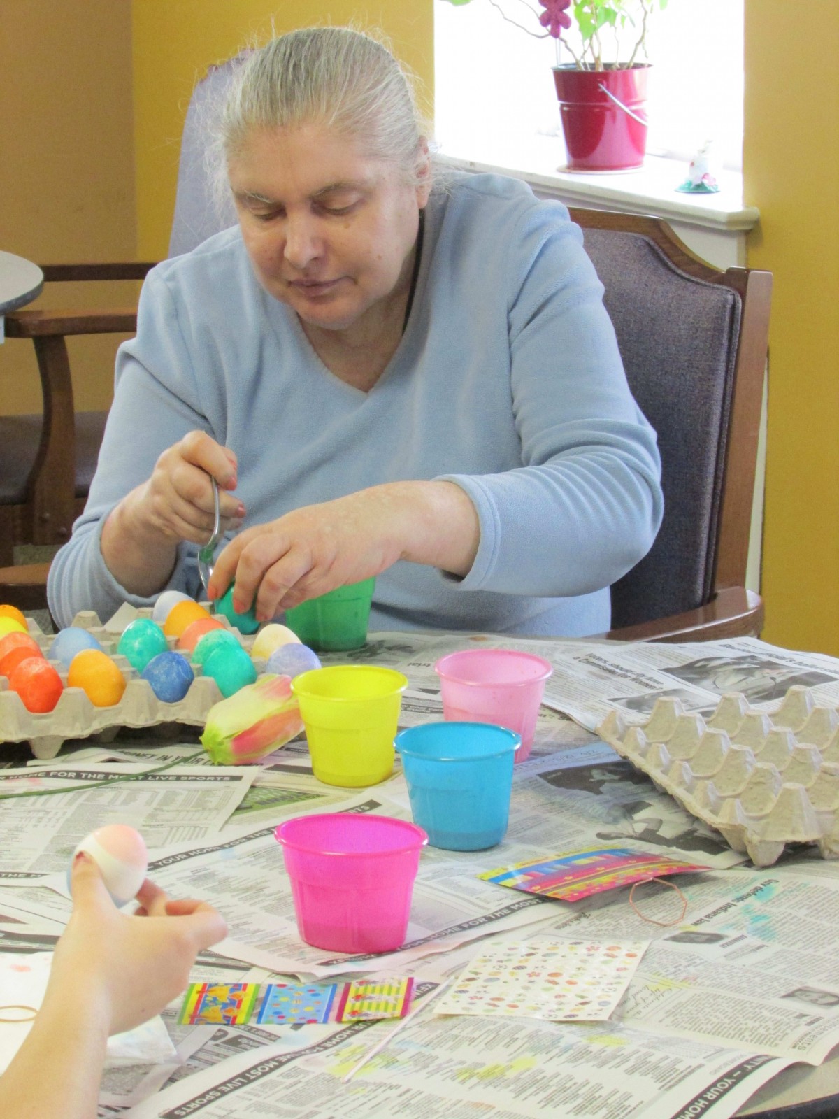 A person engaged in egg decorating, surrounded by colorful cups and newspapers on a table.