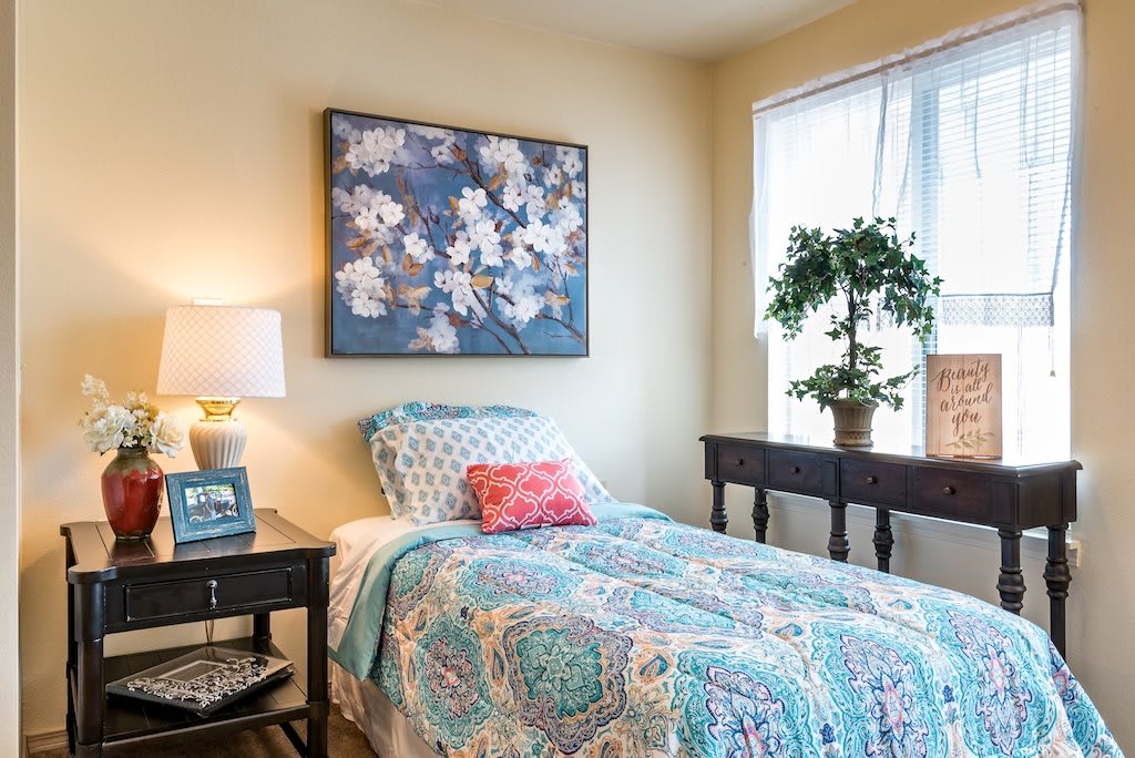 A cozy bedroom with a single bed adorned with colorful bedding, a nightstand with a lamp, floral artwork on the wall, and a potted plant by the window.