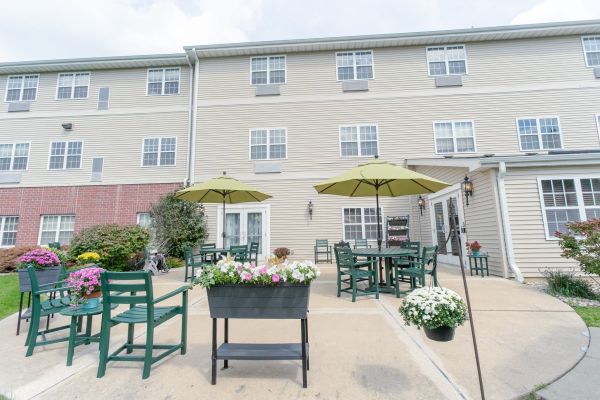 A cozy outdoor patio area with tables and chairs, featuring green umbrellas and flower decorations in front of a multi-story building.