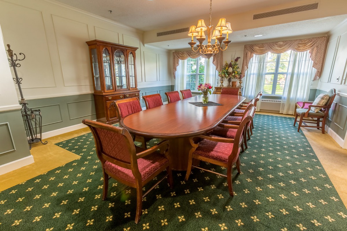 A traditional dining room with an oval table and red-upholstered chairs, illuminated by a chandelier and natural light from the windows.