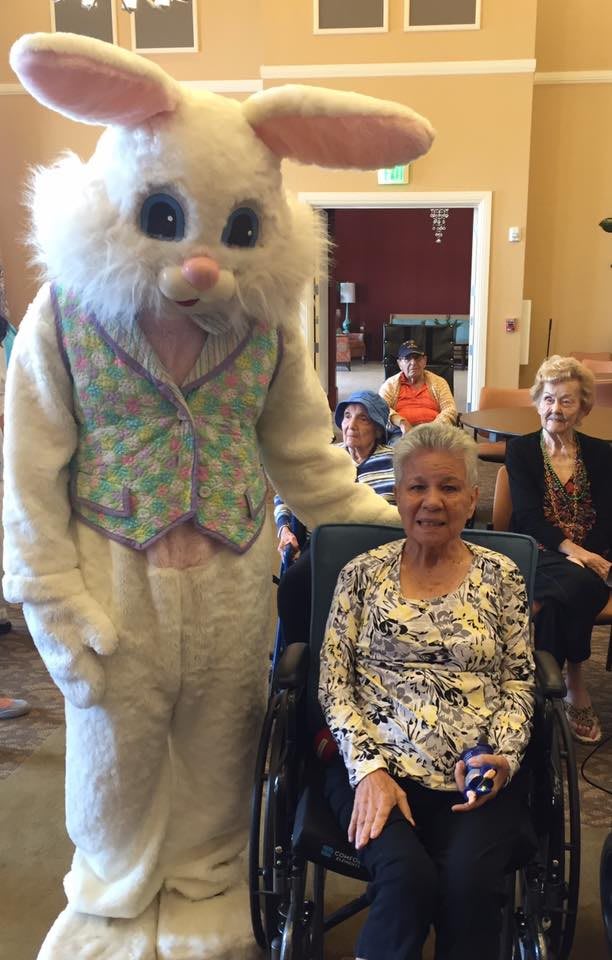 A person in an Easter bunny costume poses with an elderly woman during a festive gathering indoors.