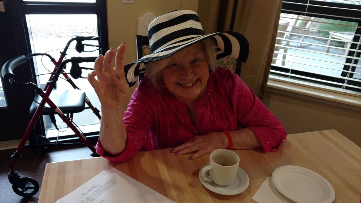 Elderly woman in a striped hat enjoying a cup of coffee and waving cheerfully.
