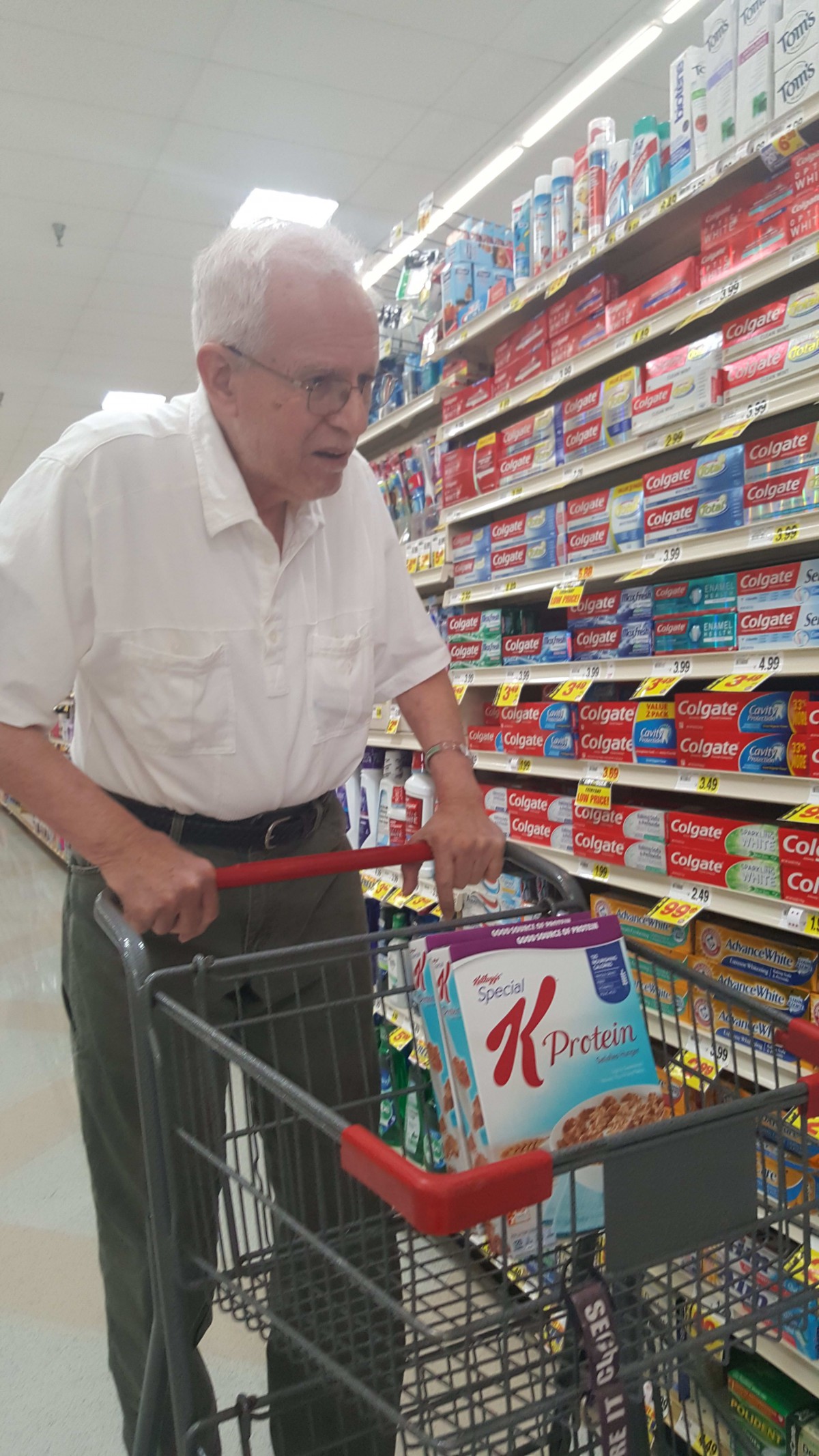 An elderly man shopping in a supermarket aisle with a grocery cart containing cereal.