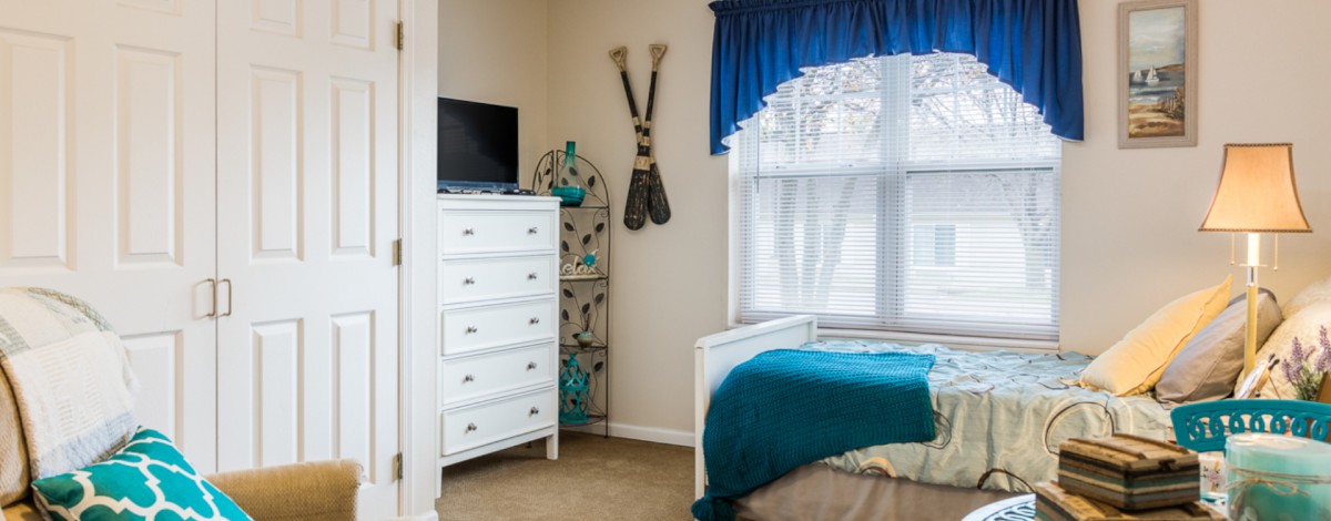 A cozy bedroom featuring a white dresser with a television on top, blue-accented decor, and a bed near a window with blue curtains.