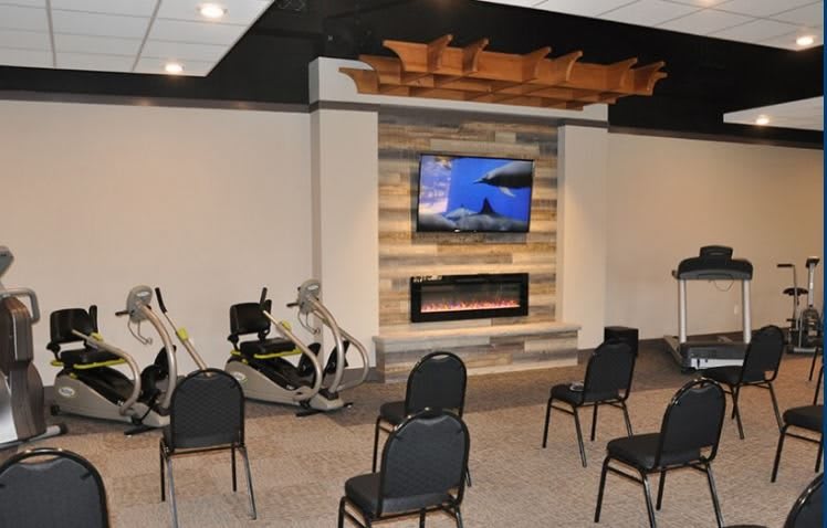 A fitness room with exercise equipment, chairs facing a television above a fireplace.