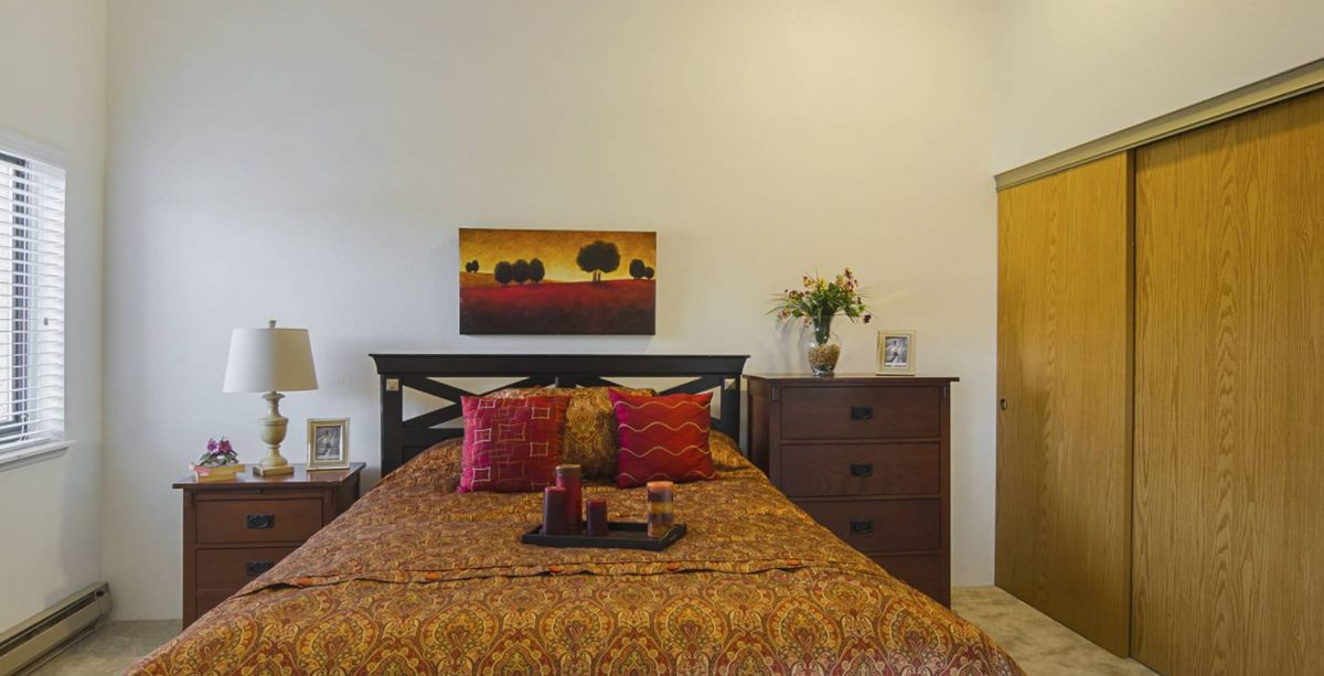 A cozy bedroom with warm-toned bedding and vibrant cushions, featuring a landscape painting above the bed and a dresser adorned with a vase of flowers.